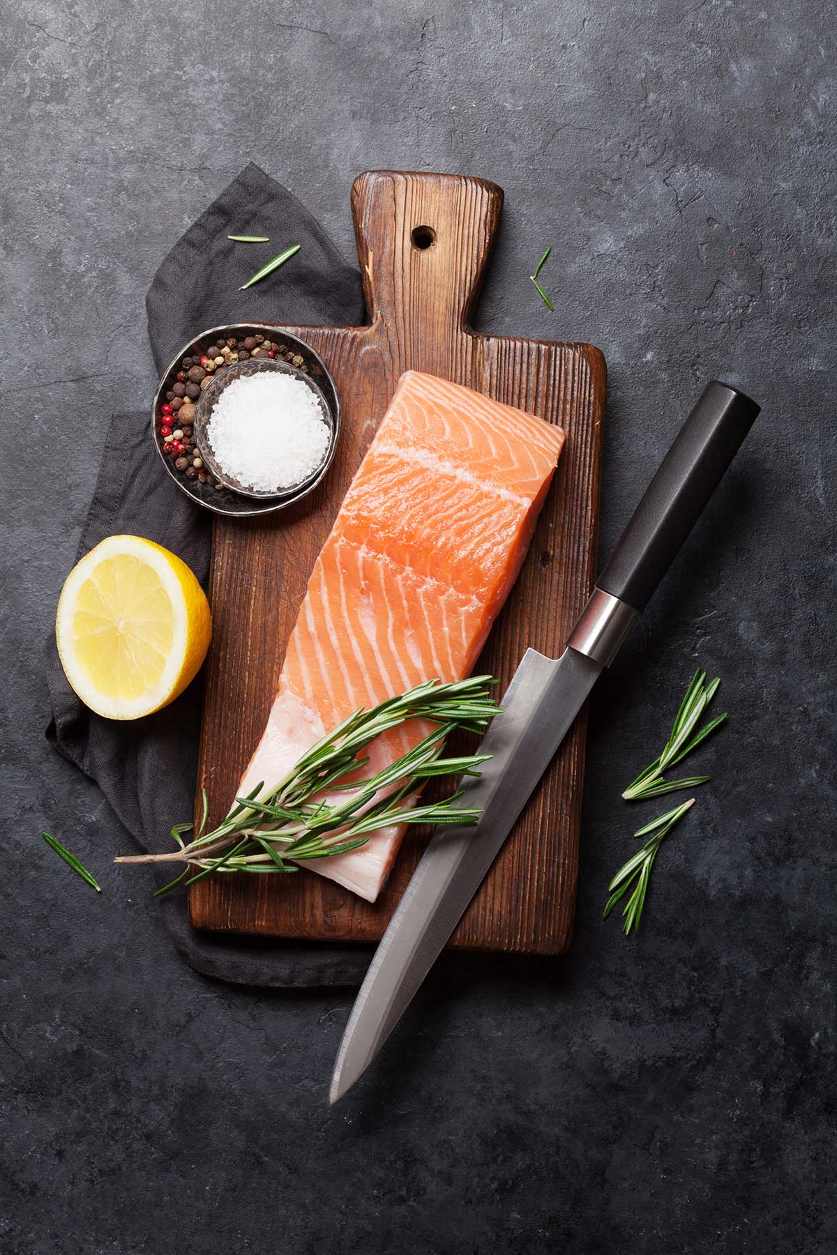 A chef's knife beside a salmon fillet, lemon and herbs on a wooden board